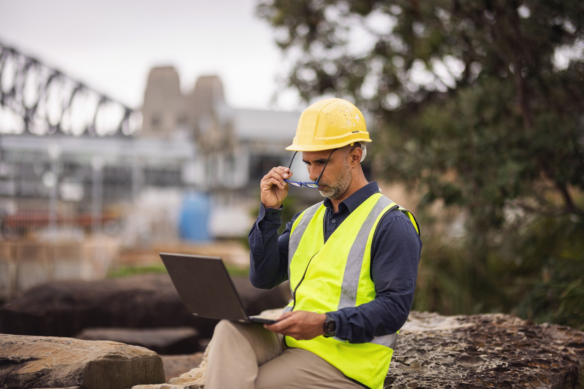 Engineer in safety vest and hard hat putting on his glasses to work on his laptop in Sydney, Australia